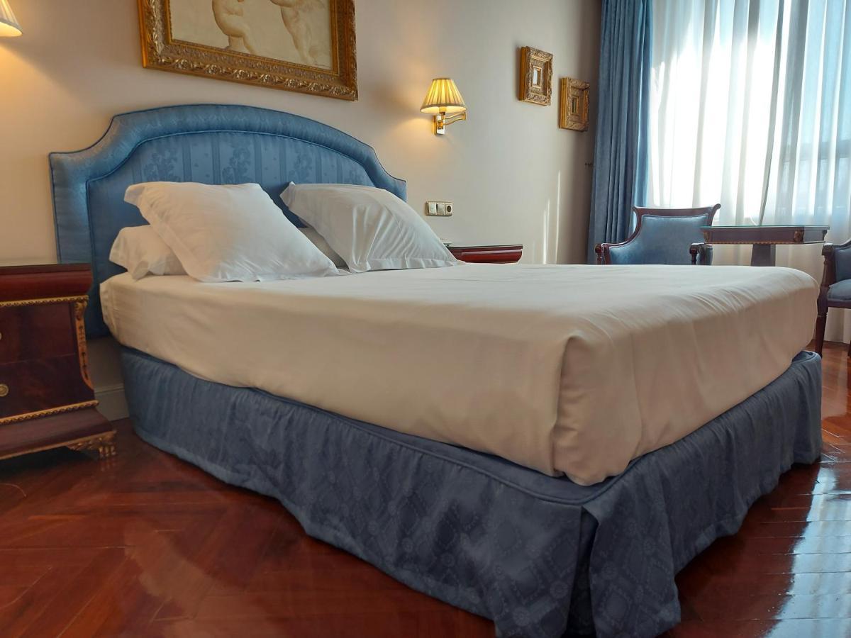 ホテル オテル アラメーダ パラス サラマンカ エクステリア 写真 The photo shows a well-furnished hotel room featuring a large bed with a light-colored mattress and decorative pillows. The bed has a soft blue headboard and a matching blue bed skirt. Beside the bed, there are bedside tables with lamps. In the backg