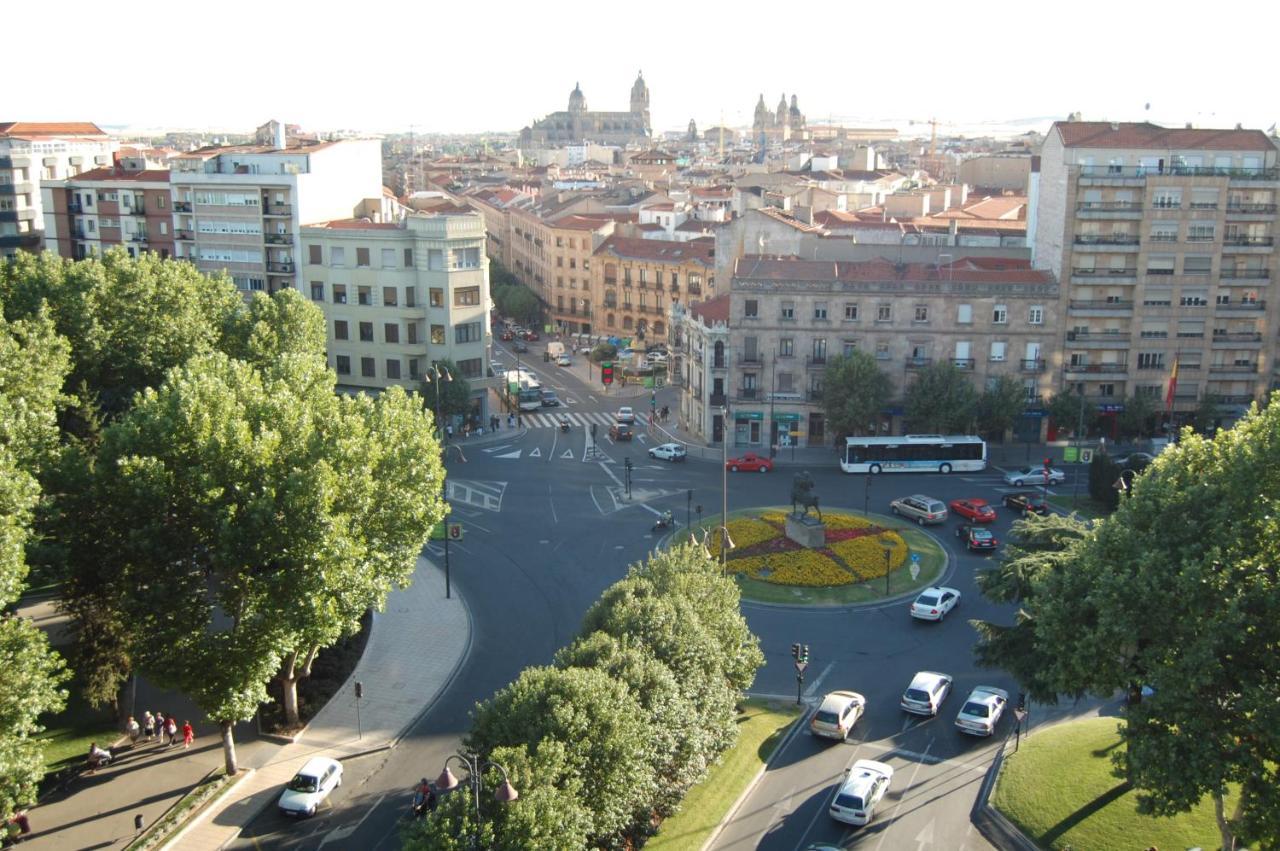 ホテル オテル アラメーダ パラス サラマンカ エクステリア 写真 The photo shows a city scene from an elevated perspective, possibly during the daytime. In the foreground, there is a roundabout with a circular flowerbed design in the center. Surrounding the roundabout, several vehicles are seen moving through the 
