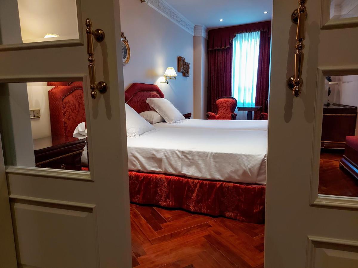 ホテル オテル アラメーダ パラス サラマンカ エクステリア 写真 The photo shows a hotel room viewed through a doorway. Inside, there is a large bed with white bedding and decorative pillows. The bed has a red upholstered headboard, and there are matching bedskirts. To the side, there's a red armchair and a window