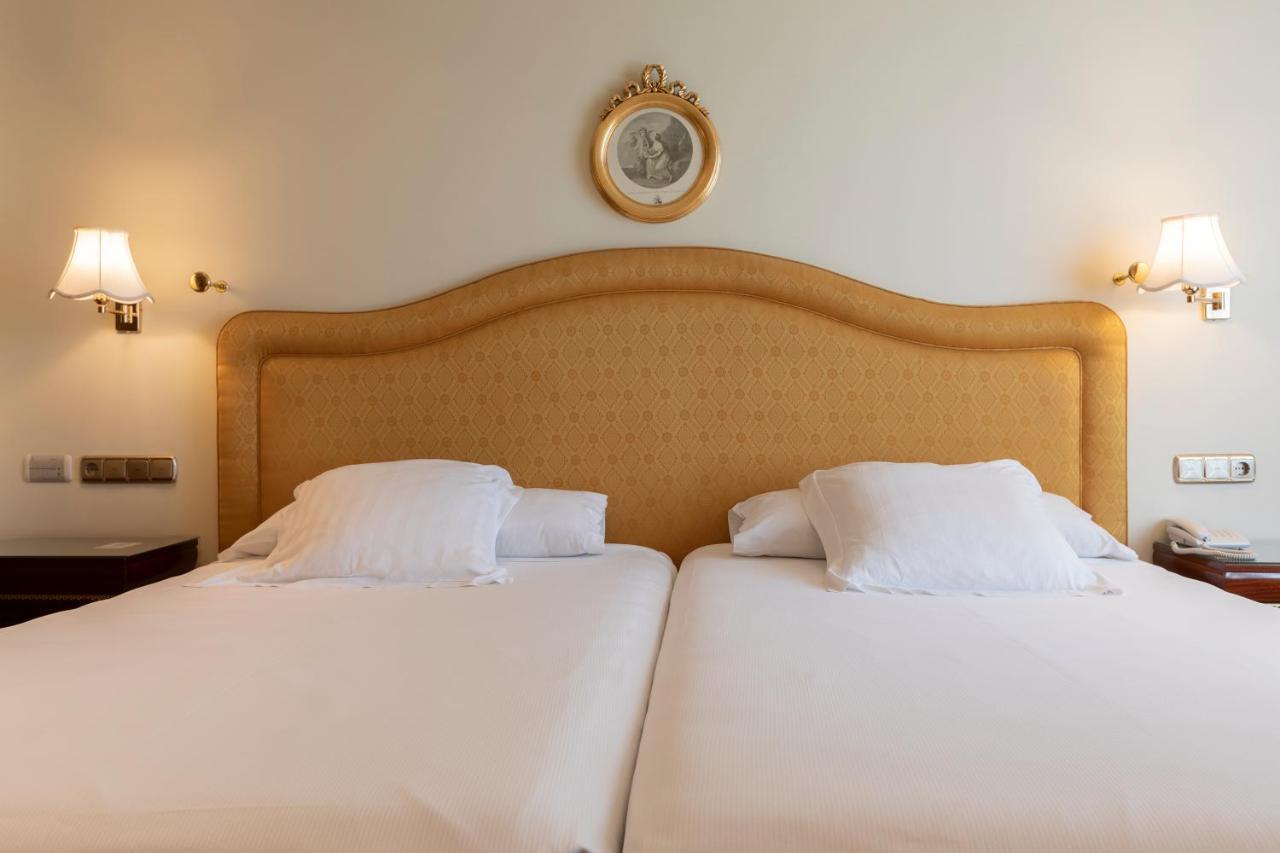ホテル オテル アラメーダ パラス サラマンカ エクステリア 写真 The photo shows a neatly arranged hotel room with two twin beds. The beds have headboards that are upholstered in a light-colored fabric, and each bed is topped with white bedding and pillows. Above the headboard, there is a decorative round wall clo