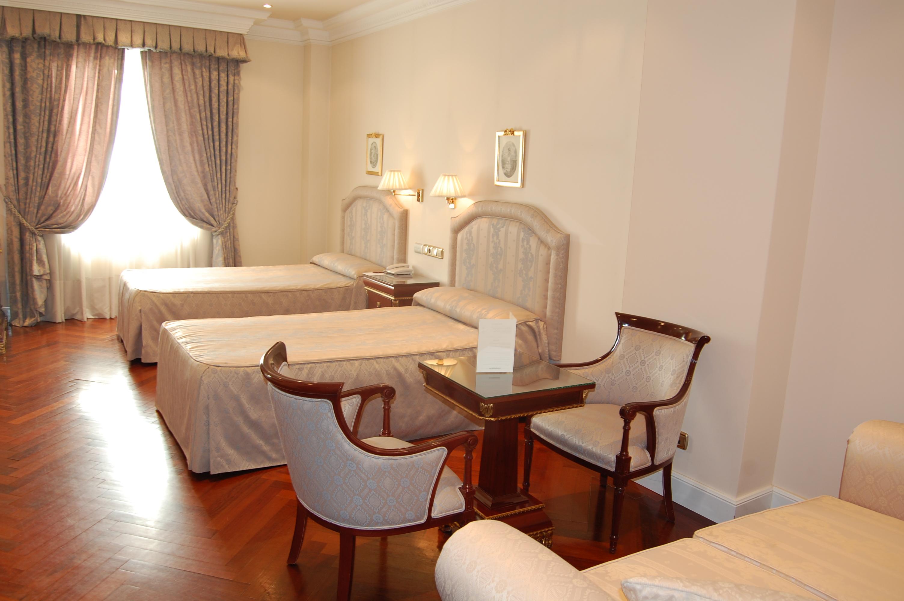 ホテル オテル アラメーダ パラス サラマンカ エクステリア 写真 The photo shows an interior of a hotel room featuring two single beds with light-colored bedspreads. There are curtains over a window allowing natural light to enter the room. A small table is positioned between two upholstered chairs, creating a coz