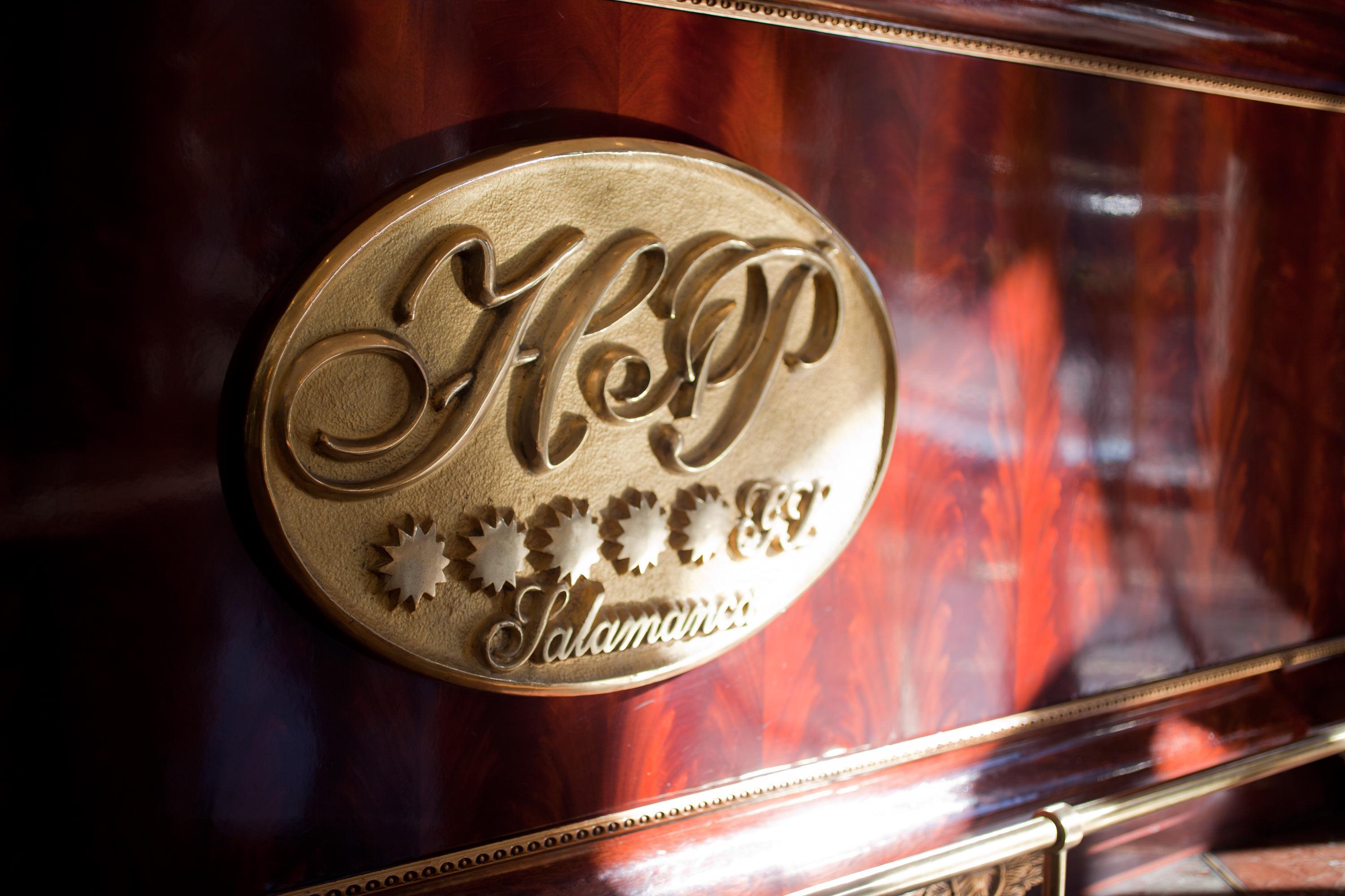 ホテル オテル アラメーダ パラス サラマンカ エクステリア 写真 The photo shows a decorative emblem with the letters "H&P" in an elegant script. The emblem appears to be made of gold or a gold-tone material and is set against a rich wooden surface, suggesting a luxurious or high-quality item. Below the letters, t