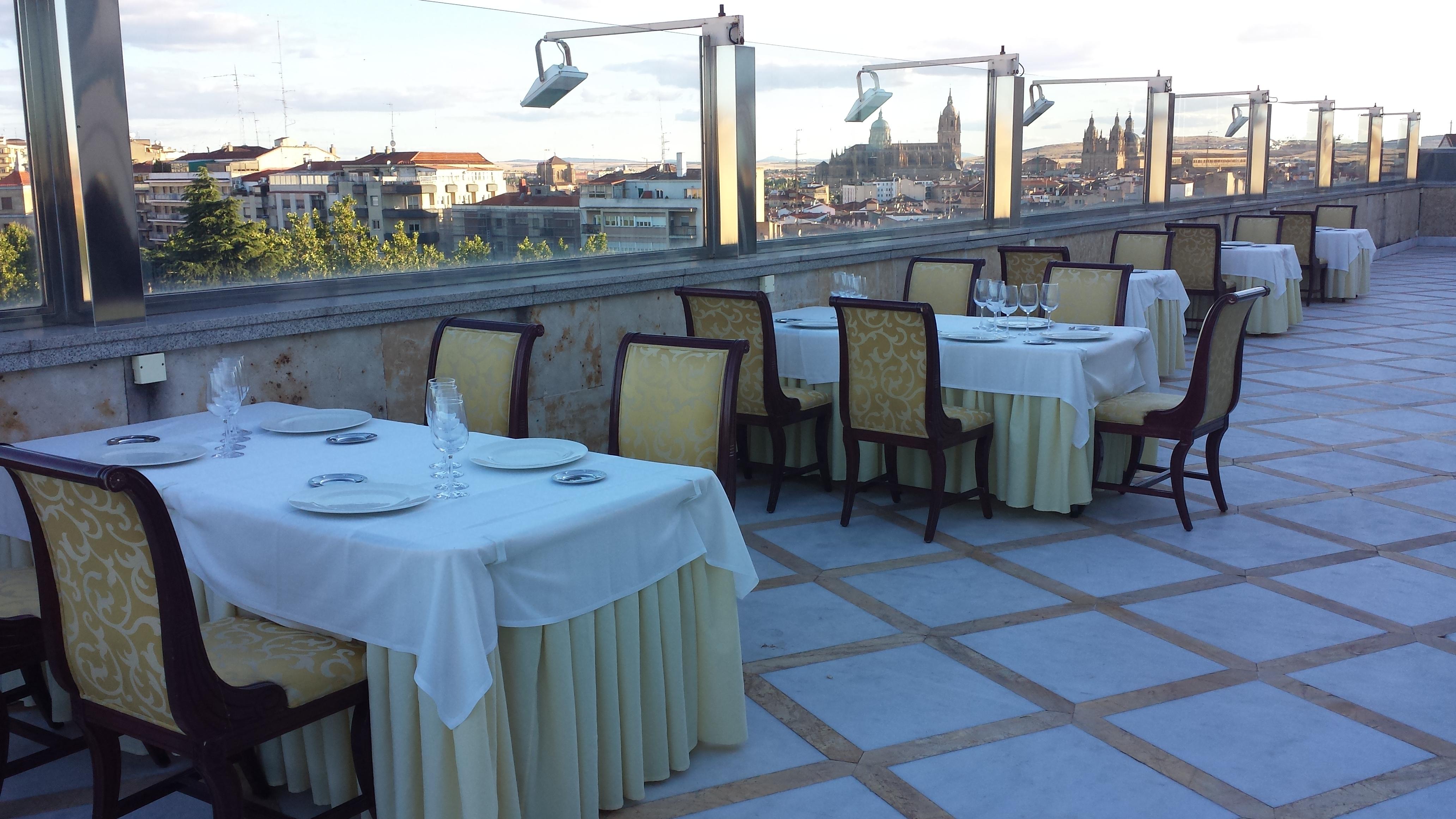 ホテル オテル アラメーダ パラス サラマンカ エクステリア 写真 The photo shows a rooftop dining area with several elegantly set tables. Each table is dressed with a white tablecloth and features chairs with ornate design. The setting offers a view of the city, visible in the background, with buildings and possib