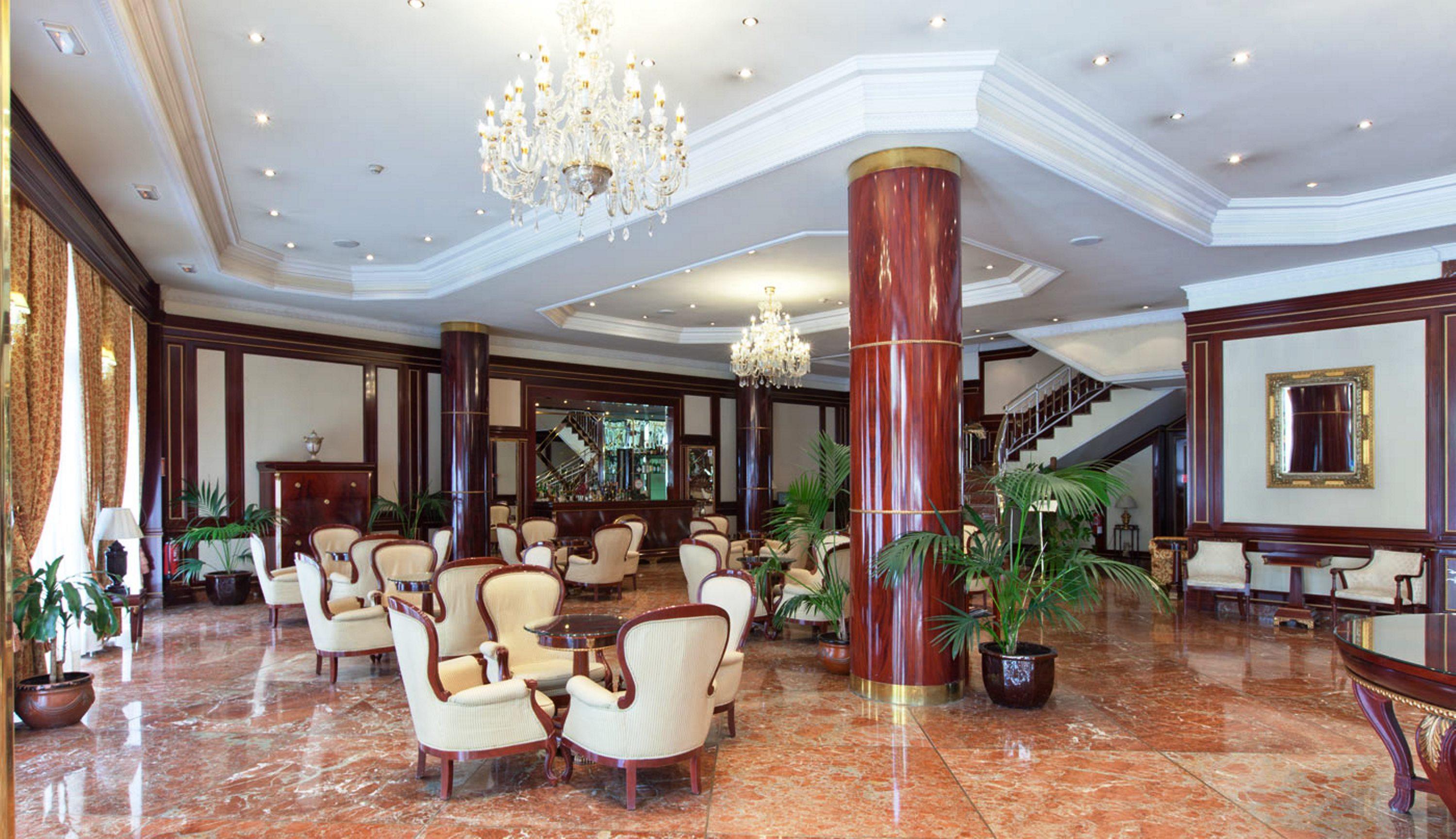 ホテル オテル アラメーダ パラス サラマンカ エクステリア 写真 The photo shows an elegant hotel lobby with a luxurious design. It features polished marble floors and rich wooden accents. There are several plush cream-colored armchairs arranged around small tables for guests to sit and relax. A grand chandelier h