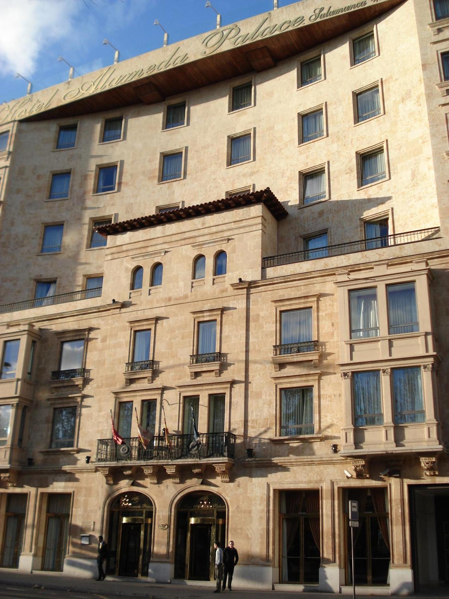 ホテル オテル アラメーダ パラス サラマンカ エクステリア 写真 The photo depicts a building with a stone facade that features a mix of architectural designs. The lower section includes several large windows framed by decorative elements, with balconies that have railings. There are also flags visible at the entr